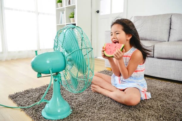 child eating watermelon with two hands. sitting in front of the electric fan.  taking a big bite delightfully.